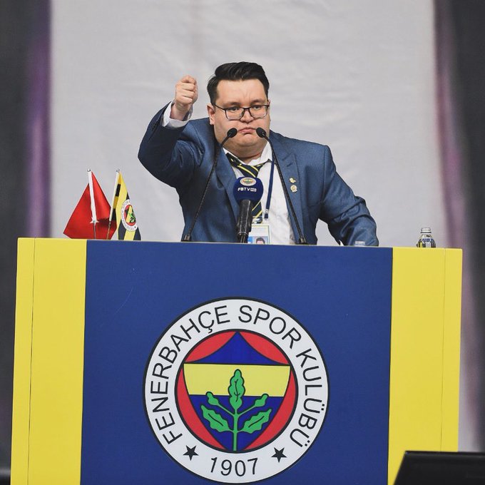 The image features a man standing at a podium with his hand raised in a thumbs-up gesture. The man appears to be wearing glasses and a suit, suggesting that he might be delivering a speech or presentation. In the background of the image, there is a banner displaying the name "Fenerbaşe Sports" along with an emblem featuring a circle and a shield. This could indicate that the event taking place is related to sports or has some connection to the Fenerbaşe Sports Foundation. The post suggests that the man being referred to might be the president of this foundation, as they are seeking a new leader for the organization.