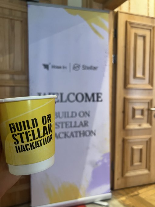 A hand holding a yellow paper cup that reads 'BUILD ON STELLAR HACKATHON' in front of a welcome banner for the same event hosted by Nx Labs and Stellar.