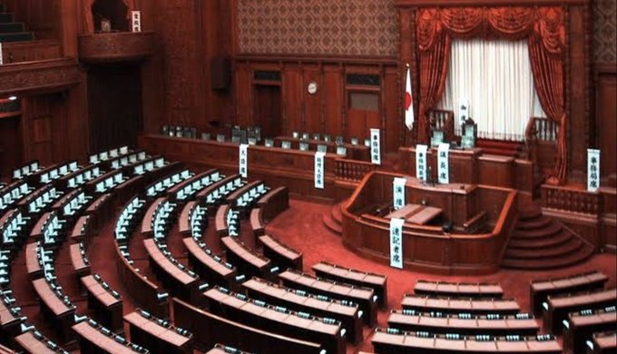 An empty Japanese parliamentary chamber featuring the national flag and labeled sections including "議長席" (Speaker's Seat) and "内閣総理大臣席" (Prime Minister's Seat), with curved rows of desks arranged in a hemicycle. The image visually represents the Japanese legislative setting where the regulatory decision to treat digital assets like stocks and bonds is being made, underscoring the post's argument about the need for traditional capital market infrastructure to support such regulation.
