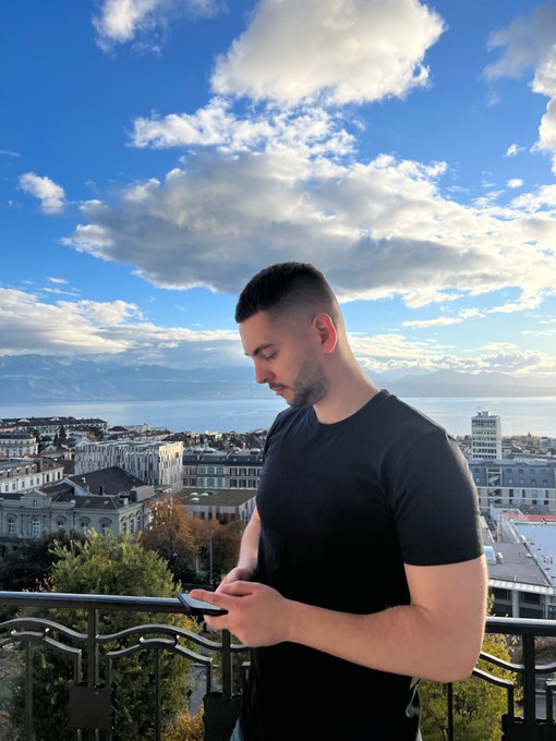 A bearded man in a black t-shirt stands on a balcony using his phone, with a cityscape, sea, and cloudy sky in the background.