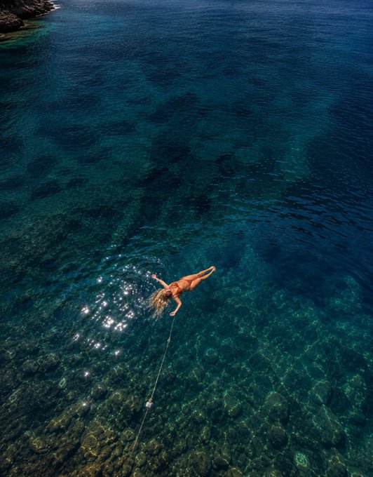 An aerial view captures a person floating on their back in strikingly clear, deep blue ocean water, tethered by a rope.
