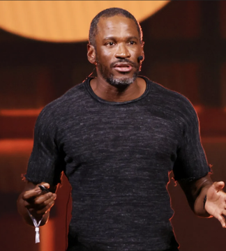 The image features a man standing against a background that seems to be a blurred square or circle with an orange hue. He appears to be wearing a black shirt. The main focus of the image is Arthur Hayes, who is holding a microphone and giving a speech. In this context, there's also a text in the image that says "Arthur Hayes is buying 1,500ETH worth $2.63M!". This information might be related to the event being discussed or another piece of Arthur Hayes' profile.