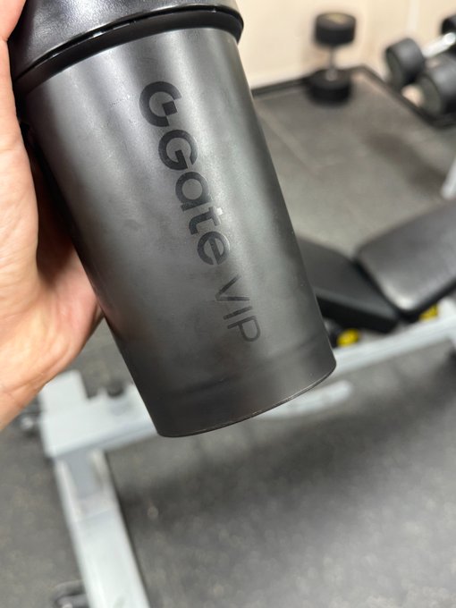 A hand holds a matte black shaker bottle branded with "G GONE VIP" in front of gym equipment.