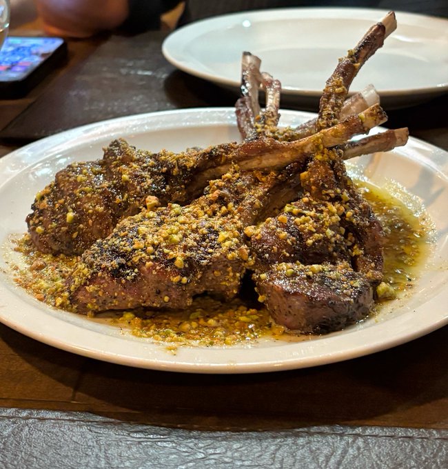 A plate of lamb chops seasoned with a crumbly herb and nut crust, served on a white plate at a restaurant table with an empty plate in the background.