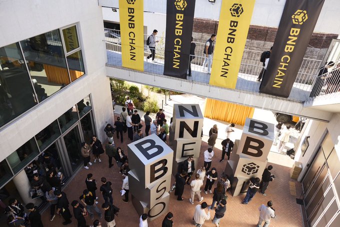 An overhead view of a crowded outdoor event with large white cube letters spelling "BNB" and "BSC" in the courtyard, surrounded by yellow "BNB CHAIN" banners.
