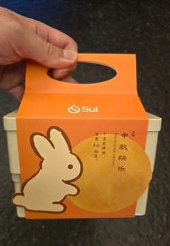 A hand holds up an orange and yellow box with a rabbit design and Japanese characters '中秋名月' (Mid-Autumn Moon), suggesting a mooncake packaging.