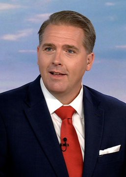 A headshot of a man in a dark navy suit, white shirt, and red tie speaking on a television broadcast.