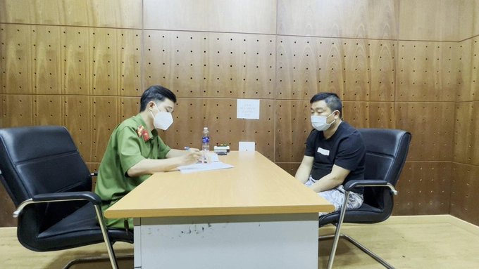 Two men wearing face masks sit across from each other at a table in a wood-paneled room; one wears a green uniform and is taking notes while the other wears a black t-shirt.