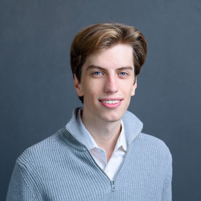 A studio portrait of a smiling young man with light brown hair, wearing a light gray sweater over a white shirt.