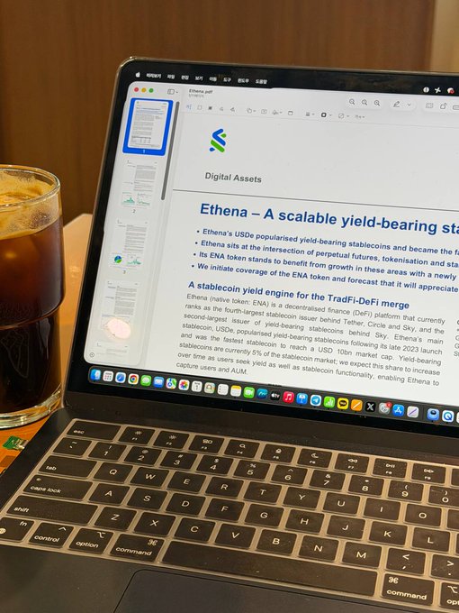 A laptop on a wooden table displays a PDF document titled 'Ethena.pdf' with the heading 'Digital Assets' and a section about 'Ethena – A scalable yield-bearing stablecoin', accompanied by a glass of iced coffee.