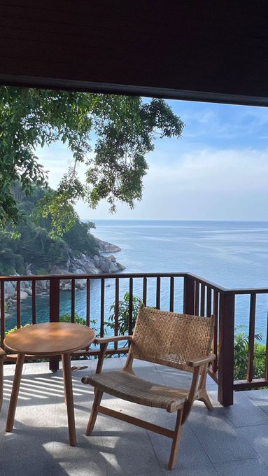 A serene balcony view overlooking the ocean, featuring a wicker chair and small round table with lush greenery framing the coastal scenery.