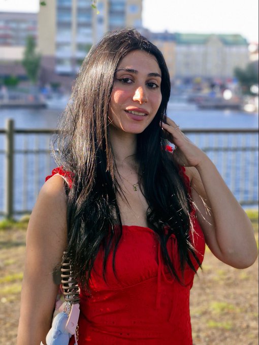 A young woman with long dark hair wearing a red dress stands outdoors near a waterfront railing, with buildings visible in the background.
