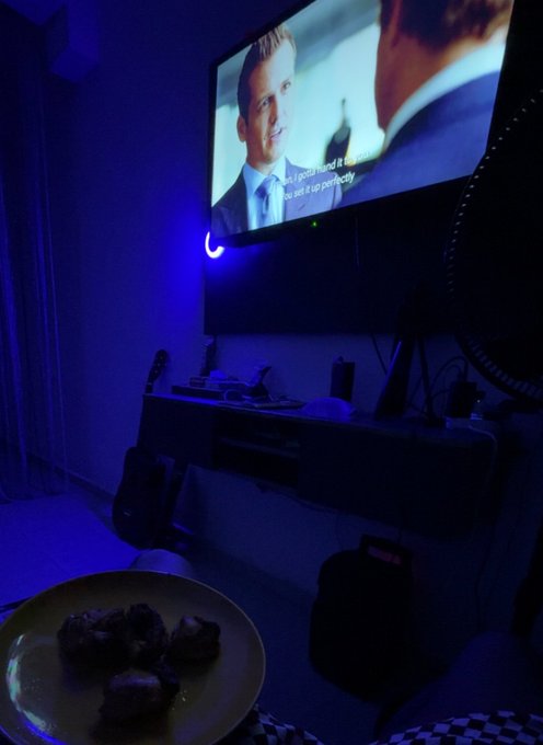 A dimly lit living room scene illuminated by blue LED ambient lighting, showing a wall-mounted TV displaying a movie scene, a floating media console with electronics, a guitar leaning against the wall, and a plate of food in the foreground.