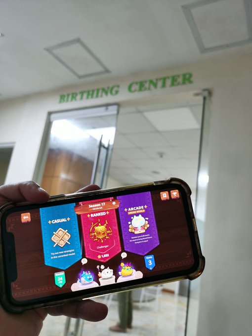 The image shows a person holding up their phone displaying a game screen with three different game options for "Getting my daily games in so my manager dont kick me out! Waiting for 5th baby to hatch. What should we name her, chat? Something @AxieInfinity related?". The person is using the phone's camera to take a picture of the game screen and is likely sharing it with others or documenting their gaming experience.