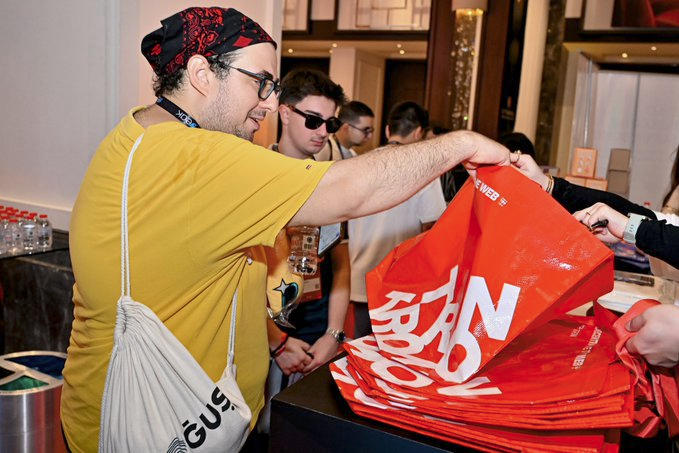 The image shows a man wearing a yellow shirt, holding an orange shopping bag with white text that says "TRON GENERATION". The scene takes place at a store or market where people are buying items from a table. There are several other people in the background who appear to be engaged in similar activities. The post about TRON generation seems to be related to this image, as it suggests that TRON is gaining popularity and becoming more accessible for everyday use.