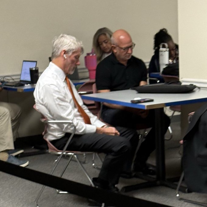 Two men in business attire — one wearing a white shirt with an orange tie and earbuds, the other in a black polo — sit attentively at a folding table alongside others in what appears to be a meeting or conference room, humorously juxtaposed with the caption as if they are "waiting for their alt to pump" at a tense board meeting.