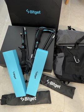 A Bitget-branded promotional setup includes a black box, blue product boxes labeled 'Bybit' and 'Bitget', a backpack, a folded camping chair, and fabric pouches, all displayed on a tiled floor.