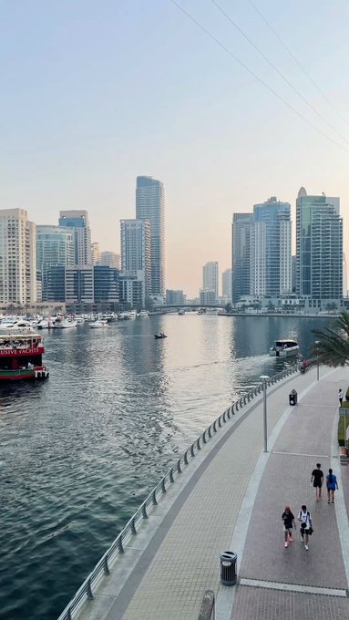 A waterfront cityscape at dusk shows modern high-rise buildings across a harbor with boats, while pedestrians walk along a paved promenade in the foreground.
