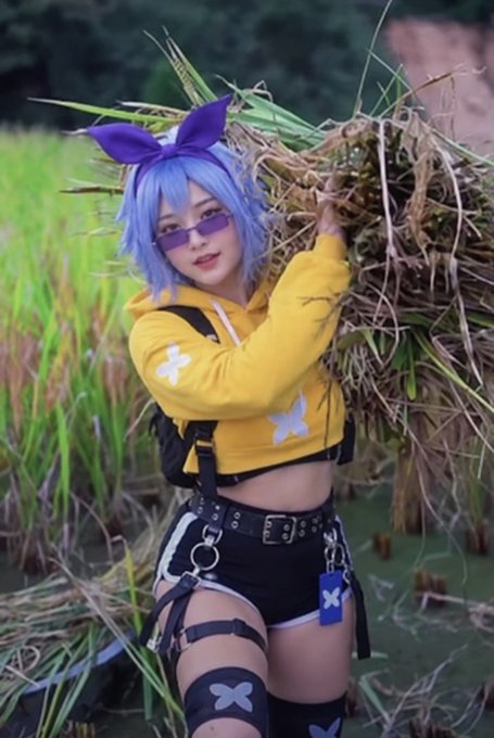 A cosplayer with blue hair and a purple bow poses outdoors with a bundle of hay, wearing a yellow hoodie with white crosses and black shorts.