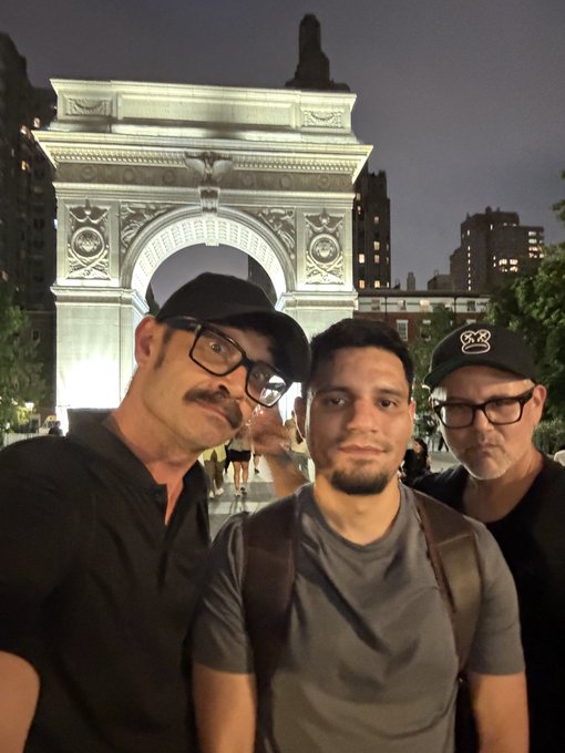 Three men pose for a selfie at night in front of the illuminated Washington Square Park arch in New York City, with the middle man wearing a backpack.