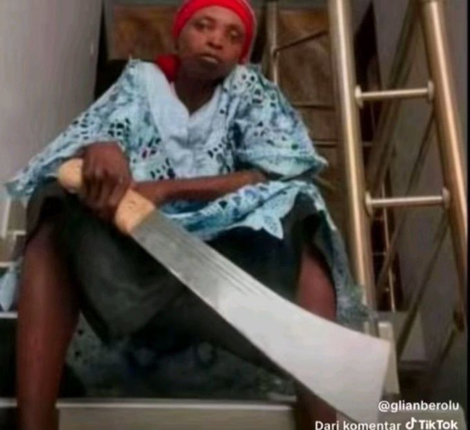 A TikTok screenshot of a person sitting on stairs holding a large machete, wearing a light blue lace top and a red head covering, with the watermark '@glianberolu' visible.