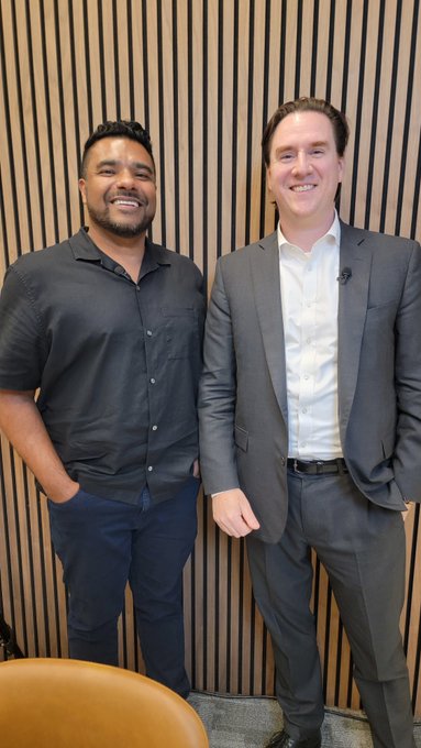 Two men smile for a photo in front of a wooden slat wall, with one wearing a black polo shirt and the other in a gray suit. The image accompanies a post celebrating a recent interview with Jim Ferraioli, Head of Crypto Research at Charles Schwab, about cryptocurrency insights.