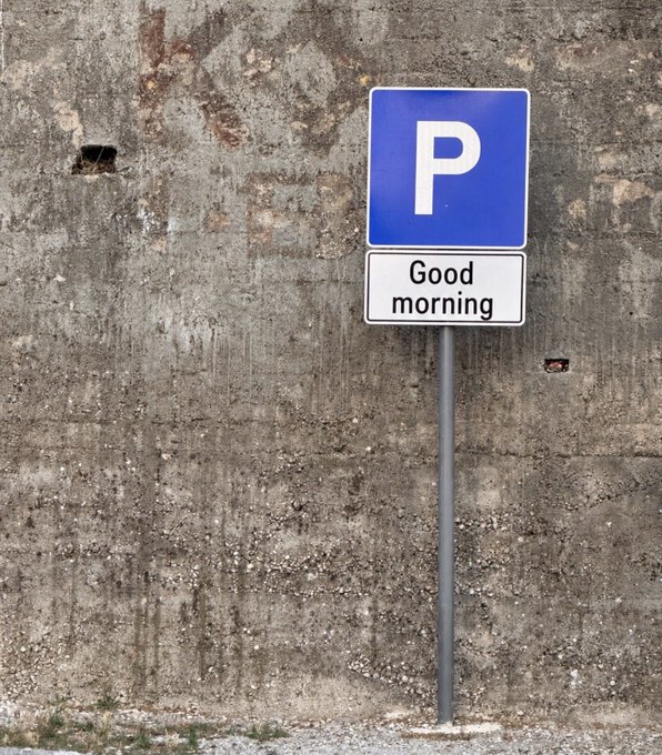 A blue parking sign with a white 'P' has a smaller white plate beneath it that humorously reads 'Good morning', mounted on a pole against a weathered concrete wall.