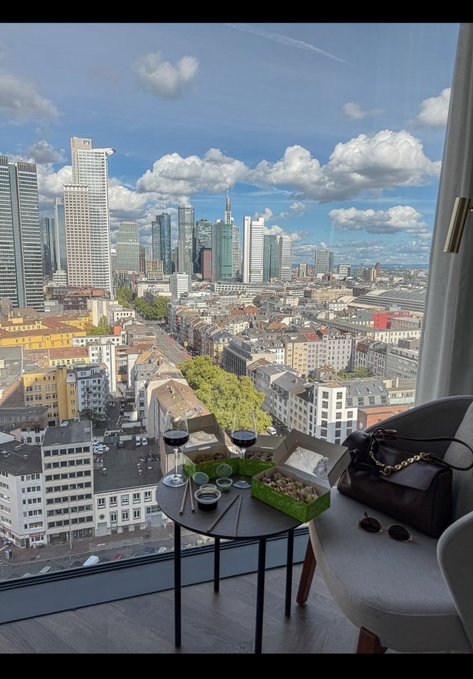 A view from a high-rise window overlooking a city skyline under a blue, cloudy sky, with a small table in the foreground holding wine glasses, sushi, and a black handbag.