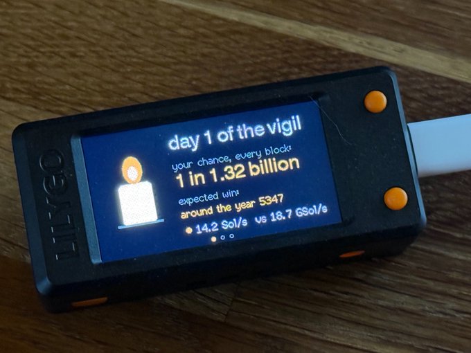 The image shows a small device with a screen displaying information about Bitcoin mining. The device has a black case and an orange border, making it stand out against the wooden background. The screen of the device displays various statistics related to Bitcoin mining, including the number of Bitcoins mined in one second (1.32 x 10^23), the estimated time needed for miners to find 1 block (5 days), and the total amount of Bitcoins expected to be mined around the year 53477 (1.52 x 10^6). The device is placed on a wooden surface, with its screen facing upwards towards the viewer.