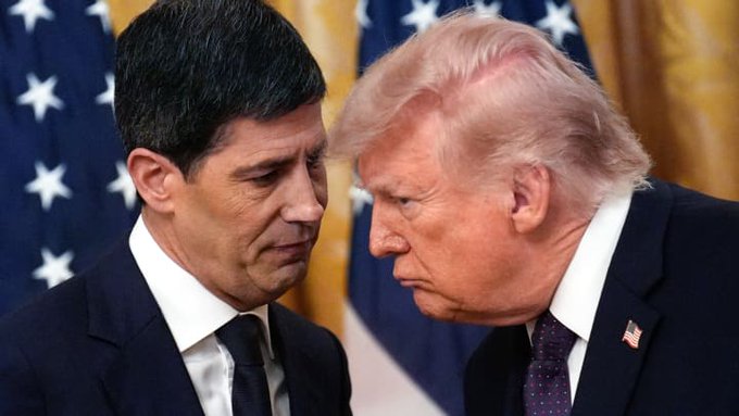 Donald Trump stands close to a dark-haired man in suits, facing him in front of U.S. flags and gold curtains.