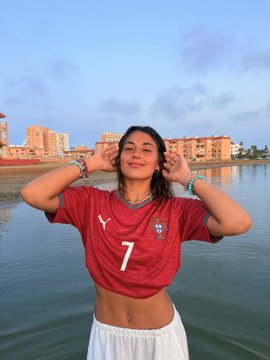 A woman wearing a red Puma jersey with the number "7" poses by the water with her hands near her ears, matching the "CrissTIAAAnoooo" celebration reference.
