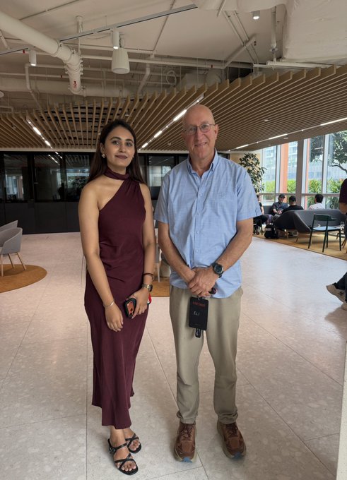 A woman in a maroon halter dress and a bald man in a light blue shirt pose together in a modern indoor venue with wooden ceiling and lounge seating.
