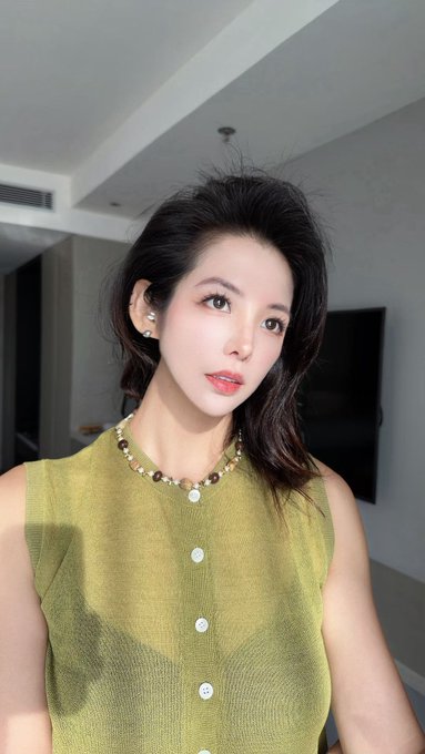 A young woman with dark hair and pearl accessories poses indoors in a soft-lit selfie, looking directly at the camera in an olive-green knit top. The image contrasts the contemplative "cash vs. Bitcoin" choice posed in the caption with a poised, reflective portrait that invites viewers to weigh their own decision between tangible wealth and digital asset.