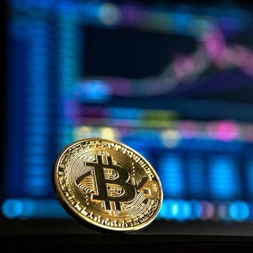 A gold physical Bitcoin coin sits in front of a blurred screen displaying a stock or cryptocurrency chart with colorful trend lines. The image visually represents the article's headline about Bitcoin's strong August performance, where it gained 24.95%—its best August since 2017—as it enters September.