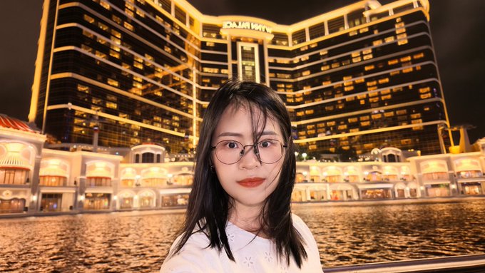 A young woman with glasses poses in front of the dazzling Wynn Palace hotel in Macau, its golden-lit facade reflected on the water and captioned "WYNN PALACE" above the entrance, perfectly capturing the opulent nighttime scene mentioned in the post.