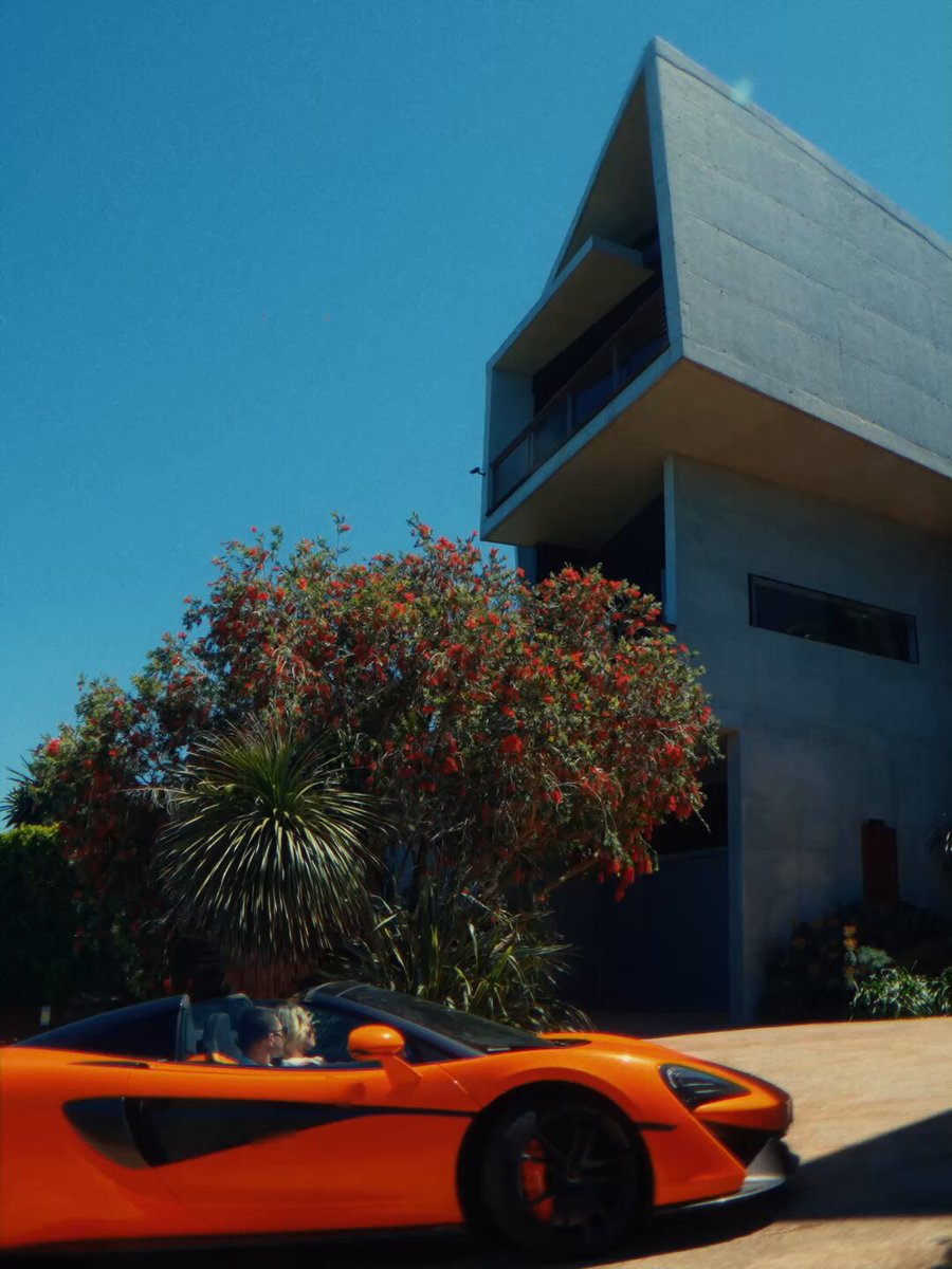 The image features a large orange sports car parked next to a tall building with an unusual triangular shape. The car appears to be a convertible, adding to its overall appeal. A person can be seen sitting inside the car, possibly admiring or enjoying the view of the surroundings. The scene suggests that the car owner is taking a break from driving and appreciating their luxurious vehicle parked outside the modern building.