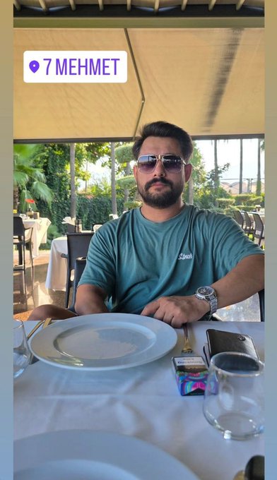A man wearing sunglasses and a teal t-shirt sitting at an outdoor restaurant table with a white plate, captioned '7 MEHMET' with a location pin.