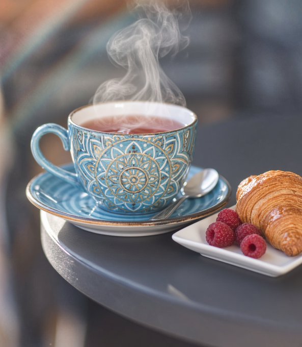A steaming cup of tea in an ornate blue mandala-patterned cup sits on a saucer beside a golden croissant and fresh raspberries on a dark table.