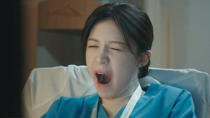 A still from a Korean drama showing a young woman in a teal hospital gown yawning widely with her eyes closed.