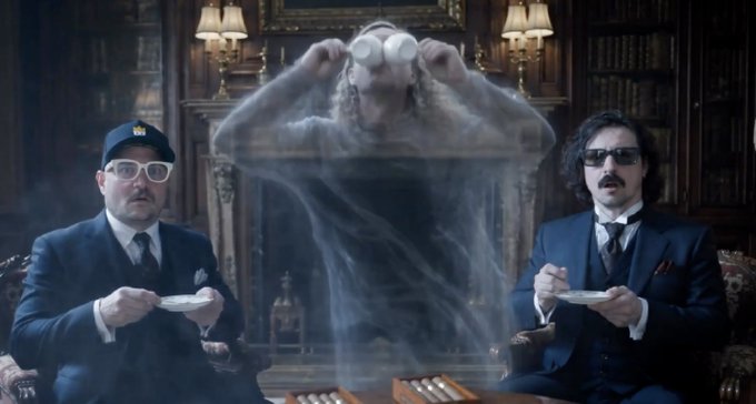 Two men in suits and sunglasses sit holding saucers while a translucent ghostly woman floats between them drinking from cups in a library setting, appearing to be a scene from a TV commercial.