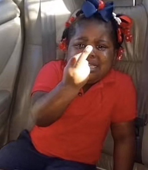 A young Black girl with red and white ribbons in her hair sits in a car seat wearing a red shirt, appearing upset and holding up her middle finger while crying.