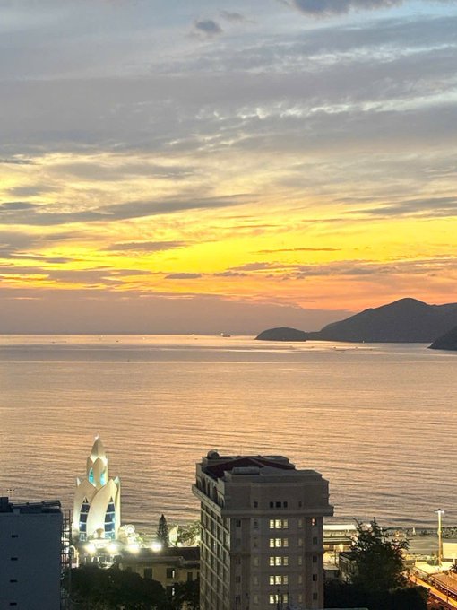 A stunning sunset over the coastal city of Vũng Tàu, with the iconic white Hải Đăng (Lighthouse) statue and its illuminated Ferris wheel standing prominently against the golden sky and calm sea, wishing everyone a warm and joyful Sunday with family.