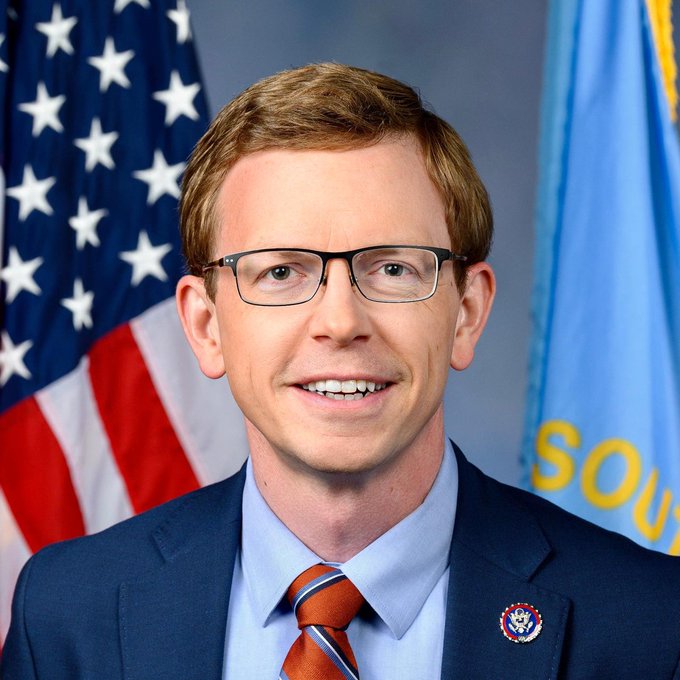 The image features a man wearing glasses, a blue suit jacket, and an orange tie with a pin that says "LATEST CLARITY Act co-sponsor Dusty Johnson says the bill must pass before another Gary Gensler-type regulator" takes control of a federal agency. The post is about the push for crypto regulatory clarity in the United States.