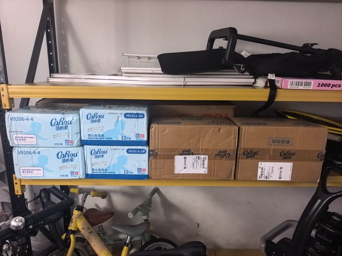 A storage shelf containing boxes of Gauyou micro-pou water filter cartridges (V9206-4-4) and other medical tissue products, with bicycles and a backpack visible below and on top.