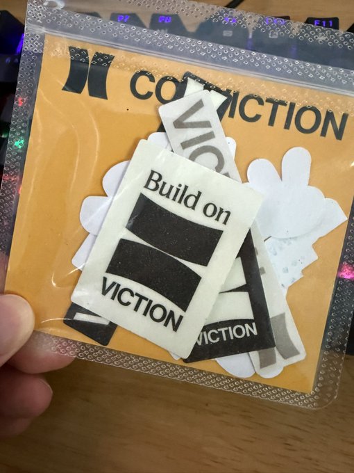 A hand holding a clear plastic package containing a sticker with the text 'Build on VICTION' on a white background, set against an orange card that reads 'VICTION CONNECTION' with a partial logo.
