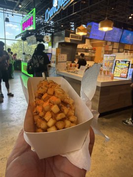 A hand holds a paper tray of loaded tater tots in what appears to be a food hall or mall setting, with a counter and customers visible in the background. The casual snack pairs with the post about @useTria's crypto cashback program, illustrating how everyday purchases like grabbing a meal can seamlessly earn Tria Points and progress toward Season 3 Mystery Boxes.