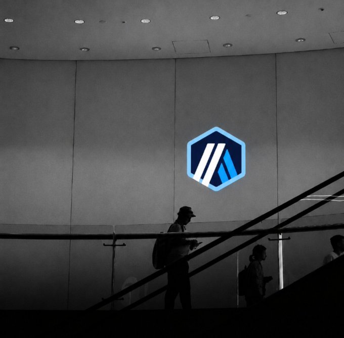 A silhouette of a person wearing a cap and backpack walking on a staircase, with another silhouetted figure visible, set against a wall displaying a projected hexagonal blue logo.