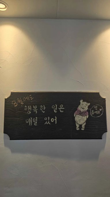 A black wooden sign on a textured wall featuring Korean text and an illustration of a sheep wearing a purple sweater, with a stamp indicating 'Newly opened.'