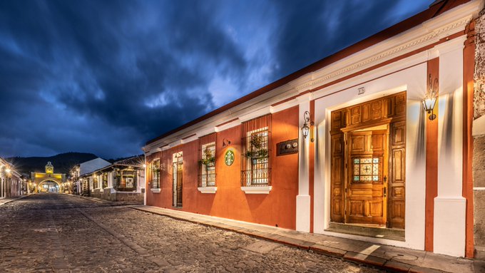 A warmly lit colonial building with a wooden door and window boxes lines a cobblestone street at dusk, illuminated by lantern-style sconces. The image visually echoes the post's message about standardized signage, as the uniform style of the building's lighting and architectural details suggests the cohesive, identifiable character that clear, consistent signage can bring to a community's cultural identity.