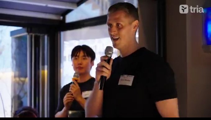 The image shows two people standing next to each other, both holding microphones. They appear to be speaking or presenting something to an audience. The background features a window that allows natural light into the room. There are also some text elements visible in the image, including the words "@useTria" and "@focus on what is in the image".